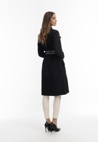 Mantou lung negru cu centură, cu textul "LOVE", contrastat de pantaloni mulți de culoare deschisă și tocuri înalte negre. Textură netedă, croială ajustată.
