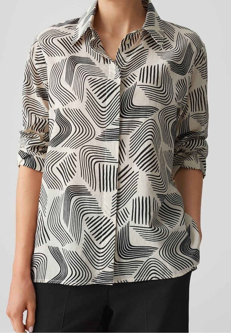 Chemise boutonnée en tissu léger, présentant un motif abstrait noir et blanc de lignes et formes courbes, avec des manches retroussées.