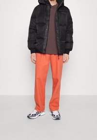 Giubbotto imbottito nero con cappuccio, pantaloni arancioni e sneakers bianco-blu. Design semplice, chiusura con cerniera, con evidente texture gonfiata.