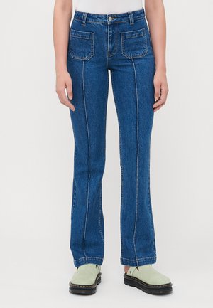 Blå denim flared jeans med hvite sømdetaljer, høy midje, to frontlommer og en lett slitt tekstur. Kombinert med grønne sko.