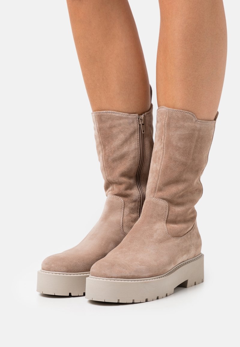 Bottes mi-mollet en daim beige avec semelles épaisses blanc cassé et fermetures éclair latérales, portées sur jambes nues sur fond blanc.