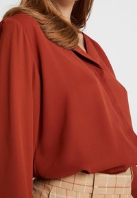 Rust-colored blouse in smooth fabric with a draped design and a notched collar, worn with tan plaid trousers featuring vertical lines.