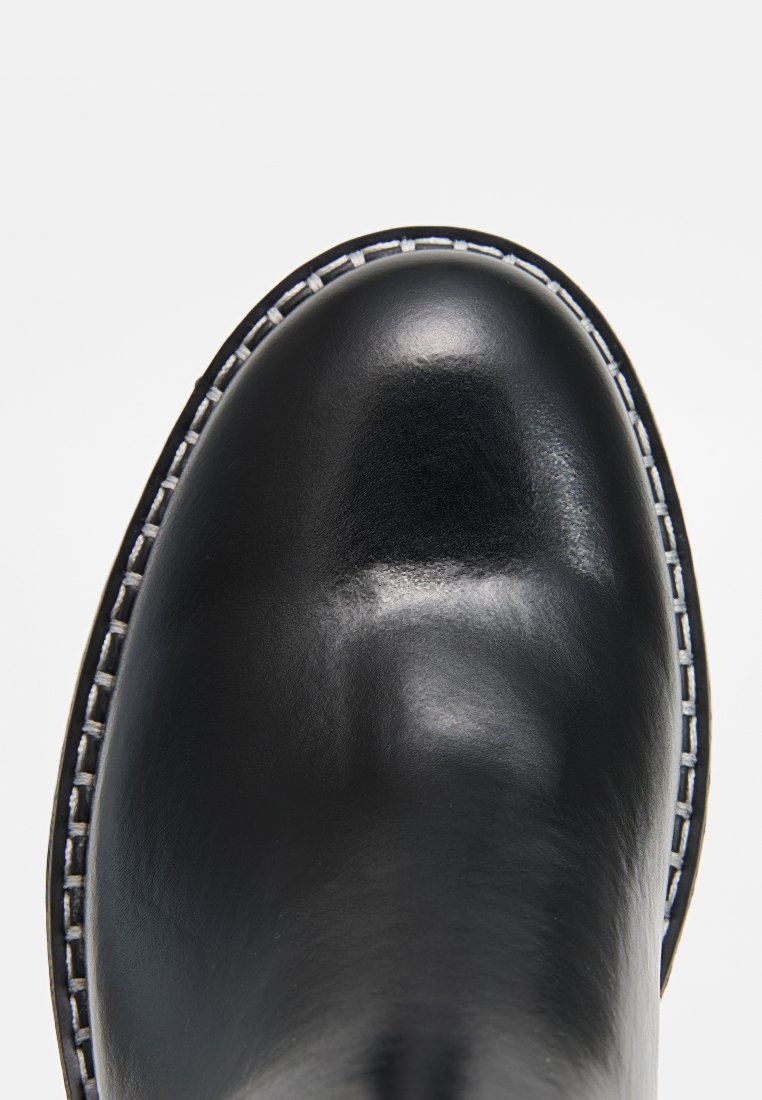 Bottine en cuir noir avec un bout arrondi, finition brillante, coutures blanches le long du bord et une texture lisse.