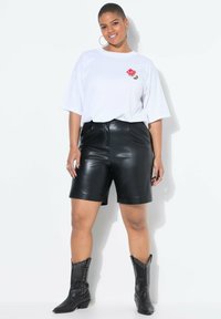 T-shirt bianco oversize con ricamo di un fiore rosa, abbinato a pantaloni shorts in finta pelle nera. Modella indossa stivali alla caviglia neri con borchie.