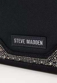 Tissu noir texturé avec un accent clouté métallique. Présente une étiquette en relief avec "STEVE MADDEN" en lettres blanches.