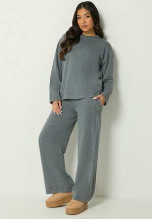 Femme aux longs cheveux foncés portant un haut ample gris foncé et un pantalon, debout avec une main dans la poche et portant des chaussures à plateforme couleur beige.