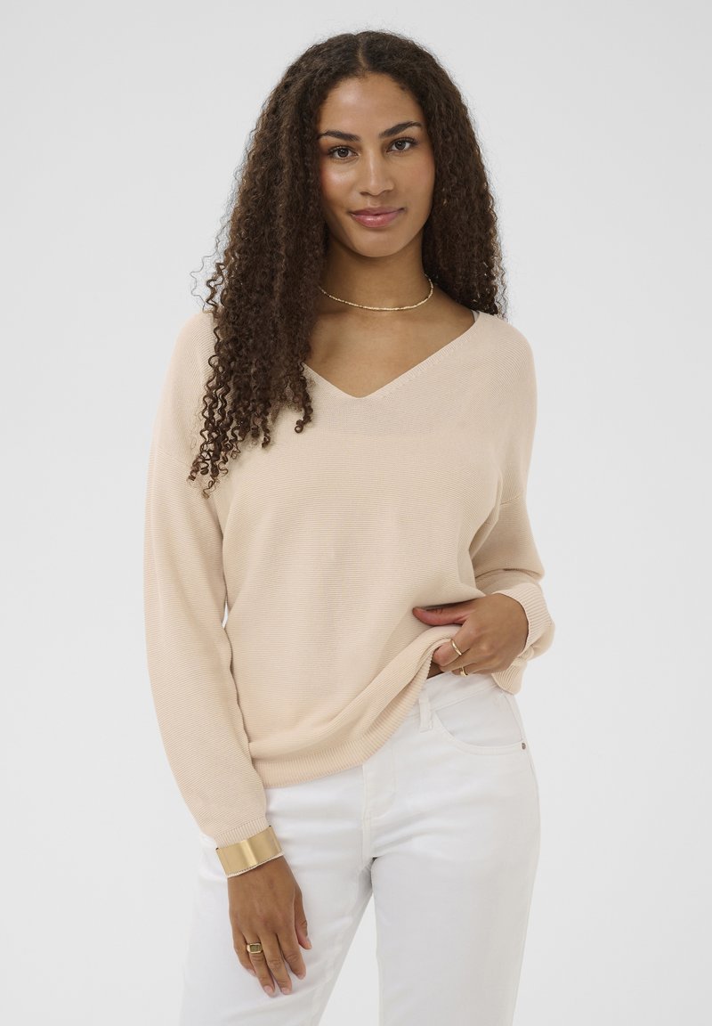 Pull beige en maille avec col en V, coupe ample et manches longues. Associé à un jean blanc et des accessoires dorés.