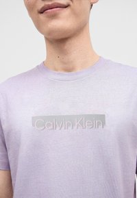 Άνδρας που φοράει ανοιχτό μωβ μπλουζάκι Calvin Klein με λογότυπο σε ανοιχτό αντιθετικό χρώμα στο στήθος, εμφανιζόμενος από τον λαιμό μέχρι το μέσο του κορμού.