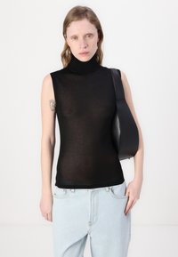 Svart ermeløs turtleneck topp i gjennomsiktig materiale, kombinert med lyseblå høytlivsjeans og en svart skulderveske. Subtil tatovering på armen.