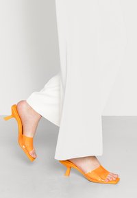Sandálias de salto com transparência laranja, com design de bico aberto e salto bloco. O material parece brilhante, com uma textura suave.