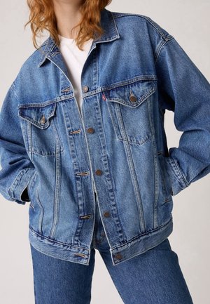 Personne portant une veste en jean bleue sur une chemise blanche avec un jean bleu assorti, debout les mains dans les poches de la veste.