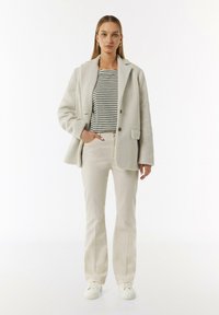 Lichtgrijze, gestructureerde, oversized jas met geknipte revers en twee knopen, gelaagd over een gestreept shirt en beige rechte broek. Witte sneakers.
