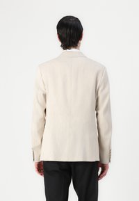 Blazer beige realizzato in tessuto testurizzato, con uno spacco singolo, revers a punta e polsini con tre bottoni. Vista da dietro, abbinato a pantaloni scuri.