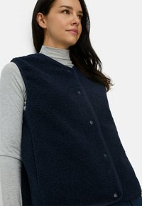 Navyblauwe fleece vest met een zachte textuur, met een ronde halslijn en een voormoet sluiting. Draagt over een lichtgrijs langemouwen top.
