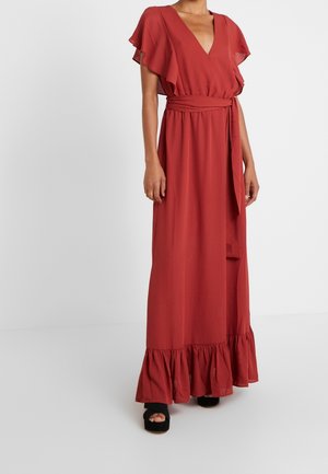 Kobieta ubrana w maxi sukienkę w kolorze rdzawej czerwieni z dekoltem w serek, z falbanami na rękawach, wiązaniem w talii, falbanką na dole oraz czarne sandały na platformie na białym tle.