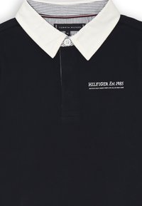 Polo navy con colletto bianco, caratterizzato da un bottone centrale e un logo ricamato "HILFIGER Est. 1985" sul lato sinistro. Tessuto in cotone.