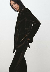 Blazer de terciopelo marrón oscuro con un dise�ño de doble botonadura, botones dorados y un corte holgado, combinado con pantalones a juego sobre un fondo claro.