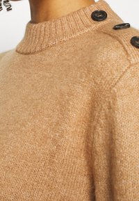 Maglione a maglia color cammello con colletto alto a coste e tre bottoni scuri sulla spalla, indossato da una persona con capelli ricci.