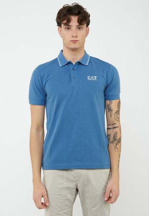 Joven con cabello castaño que lleva una camisa polo azul EA7 y pantalones claros, mostrando su antebrazo derecho tatuado sobre un fondo blanco liso.