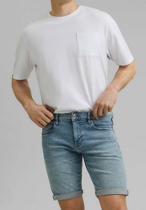 Mann trägt ein schlichtes weißes T-Shirt mit kurzen Ärmeln und Tasche sowie hellblaue, hochgekrempelte Jeansshorts, Hände justieren den Hosenbund.