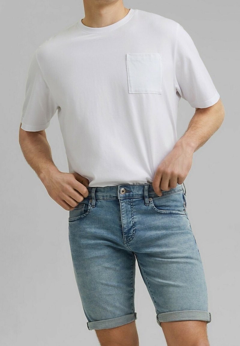 Homme portant un t-shirt blanc uni à manches courtes avec poche et un short en denim bleu clair retroussé, les mains ajustant la taille.