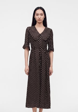 Femme aux longs cheveux noirs porte une robe midi marron à pois blancs, un grand col arrondi et des manches trois-quarts.