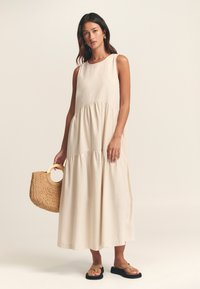 Dlhé bezrukávové šaty zo svetlo béžovej tkaniny s viacerými vrstvami. Nosené s čiernymi sandálmi, držiaca pletenú slamennú tašku s kruhovým uchom.