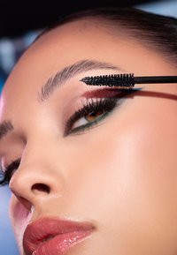 Primo piano di un volto che applica mascara nero con un pennello curvo. Presenta un trucco occhi audace con eyeliner verde e labbra lucide. Texture della pelle liscia.
