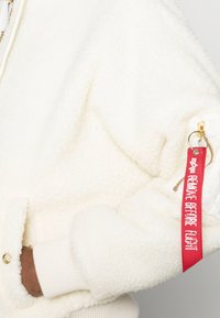 Valkoinen fleece-takki, jossa on teksturoitu pinta, ribbaantuneet hihansuut ja punainen "REMOVE BEFORE FLIGHT" -tunnus, joka on kiinnitetty vetoketjuun.