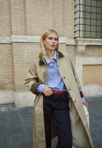 Bež baloner s smeđim ovratnikom, plava prugasta košulja, crveni remen i tamne traper hlače. Model pozira ispred teksturiranog kamenog zida.