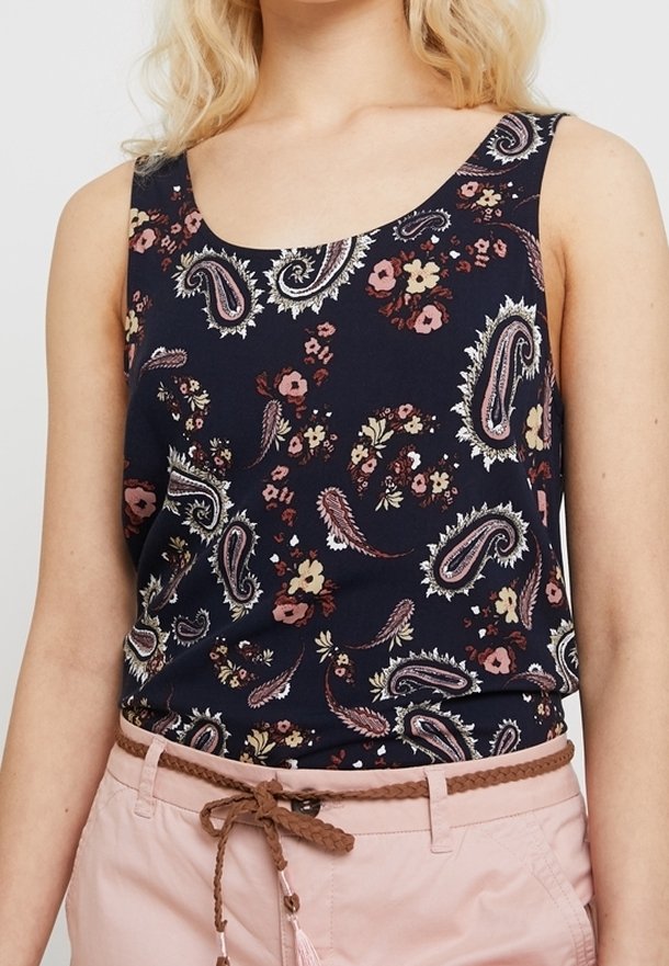 Femme portant un haut sans manches bleu marine avec des motifs cachemire et floraux roses et beiges, assorti d’un pantalon rose clair et d’une ceinture tressée marron.