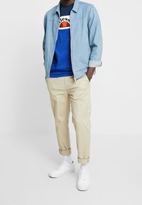 Muž v světle modré denimové bundě, modrém tričku s potiskem, béžových kalhotách s ohrnutými lemy, bílých ponožkách a bílých teniskách, stojí s rukama v kapsách.