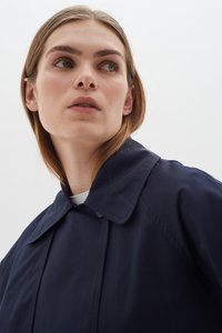 Tumši zila lietussargu mēteli ar lielu apkakli, izgatavots no gludas auduma, ar apakšmali un pogu detaļām, uzvilkts virs baltas blūzes.