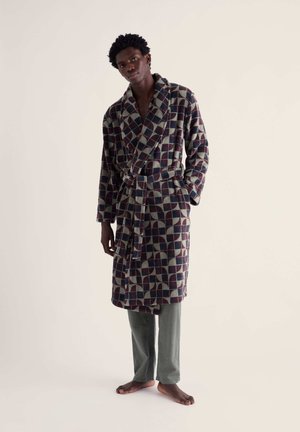 Homme debout pieds nus portant un peignoir à motifs par-dessus un pantalon gris sur un fond clair uni.