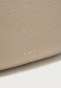 Κοντινή εικόνα από μπεζ υφή δέρματος με μικρό χρυσό ανάγλυφο λογότυπο "FURLA" σε μια μαλακή καμπυλωτή επιφάνεια.