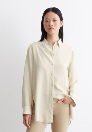 Camisa de botones crema con cuello, mangas largas y corte relajado, hecha de un tejido suave. Detalles de abertura lateral notables.