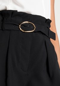 Černý látkový pásek s velkou, nepravidelnou zlatou sponou, zdobený skladováním na tmavých kalhotách, zajištěný černými knoflíky.