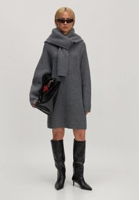 Graues ribbeliges Strickkleid im Oversized-Stil mit einem passenden Schal, kombiniert mit eleganten schwarzen knielangen Stiefeln und einer glänzenden schwarzen Handtasche.