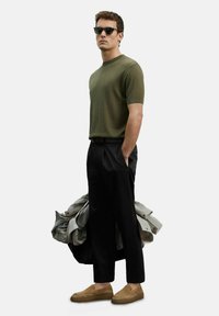 Olijfgroene geribbelde korte mouwen shirt, zwarte broek, bruine instappers. Houdt een lichtgrijze jas vast, met donkere zonnebril, staand op een effen achtergrond.