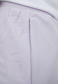 Helle lavendelfarbene Sporthose aus strukturiertem Material, mit einem durchbrochenen Logo und seitlichen Taschen, die ein glattes, flexibles Design zeigt.