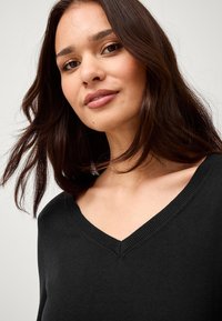 Maglione nero a V con scollatura a costine, caratterizzato da una texture morbida. I capelli sono sciolti e mossi, incorniciando il viso. Orecchino sottile visibile.