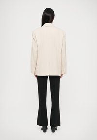Woman with long dark hair stands facing away, wearing an oversized beige blazer, black flared pants, and black heeled shoes on white background.
