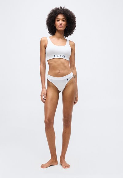 Γυναίκα όρθια ξυπόλητη, φοράει αθλητικό σουτιέν Polo σε λευκό χρώμα με το λογότυπο και ταιριαστά σλιπ, μπροστά σε απλό φωτεινό φόντο.