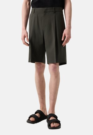Mann trägt dunkelolivfarbene, kniehohe, maßgeschneiderte Shorts und schwarze Sandalen mit breiten Riemen, steht vor einem schlichten hellen Hintergrund.