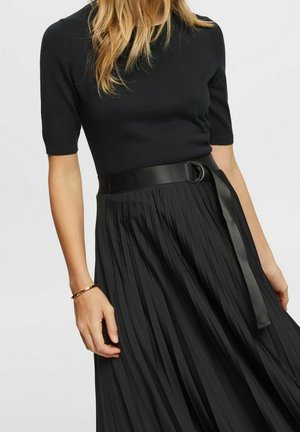 Nainen, joka pukeutunut mustaan lyhythihaiseen yläosaan, joka on tungettu korkeavyötäröiseen mustaan laskostettuun hameeseen, leveän mustan vyön ja kultaisen rannekorun kanssa.