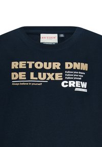Marineblauwe T-shirt met ronde hals en beige en witte opdruk. Bevat de teksten "RETROUR DNM DE LUXE" en "Always believe in yourself." Zacht materiaal.