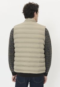 Beige pufferväst med horisontellt quiltat mönster, hög krage och ribbad nederkant, lager på lager över en mörk, strukturerad stickad tröja.