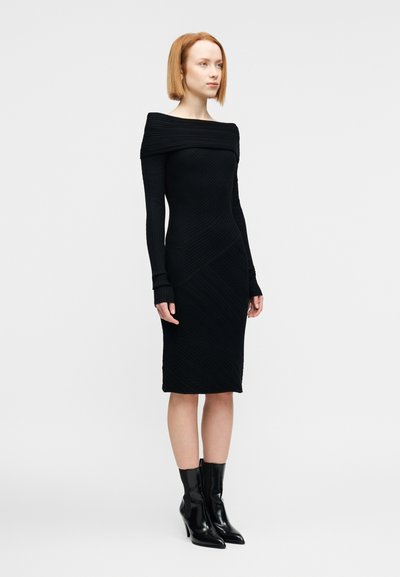 Μαύρο πλεκτό φόρεμα με ντεκολτέ που φεύγει από τους ώμους, μακριά μανίκια και διαγώνια υφασμάτινα σχέδια. Συνδυάζεται με μαύρα μποτάκια μέχρι τον αστράγαλο.