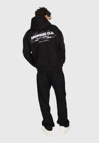 Person in einem schwarzen Hoodie mit weißem Text "Dropsize Clo." und schwarzen Hosen, stehend mit dem Rücken zu einer einfarbigen hellen Hintergrund.