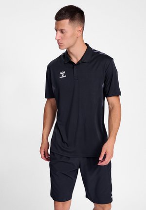 Schwarzes Polohemd mit Kragen, auf der Brust mit einem Logo versehen, kombiniert mit schwarzen Shorts. Das Hemd hat Schulterdetails in einem helleren Farbton.